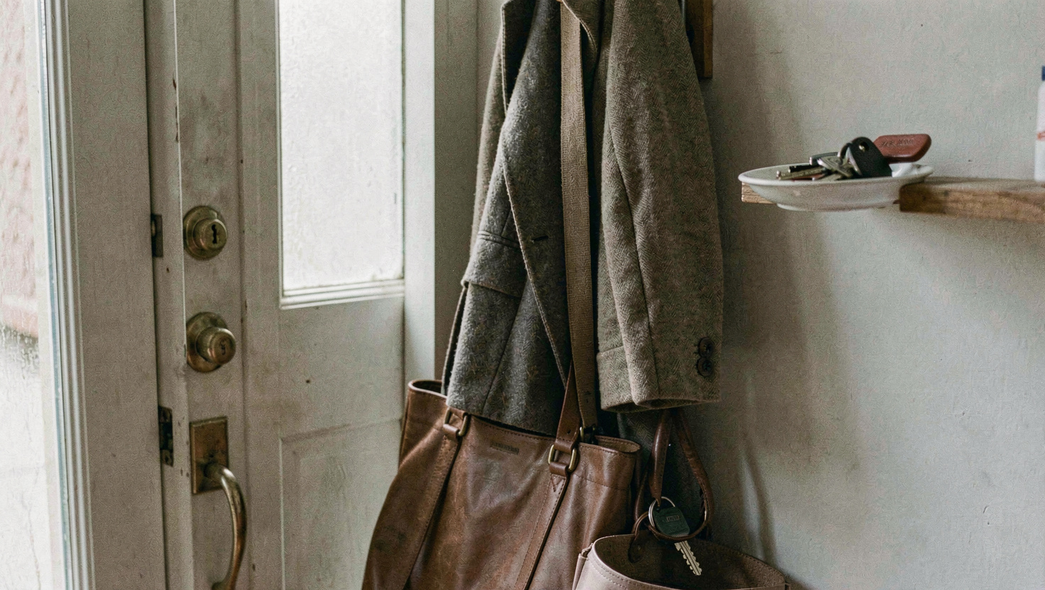 A coat hanging on a hook by a front door with a bag propped underneath it and a set of keys in a small dish on a shelf