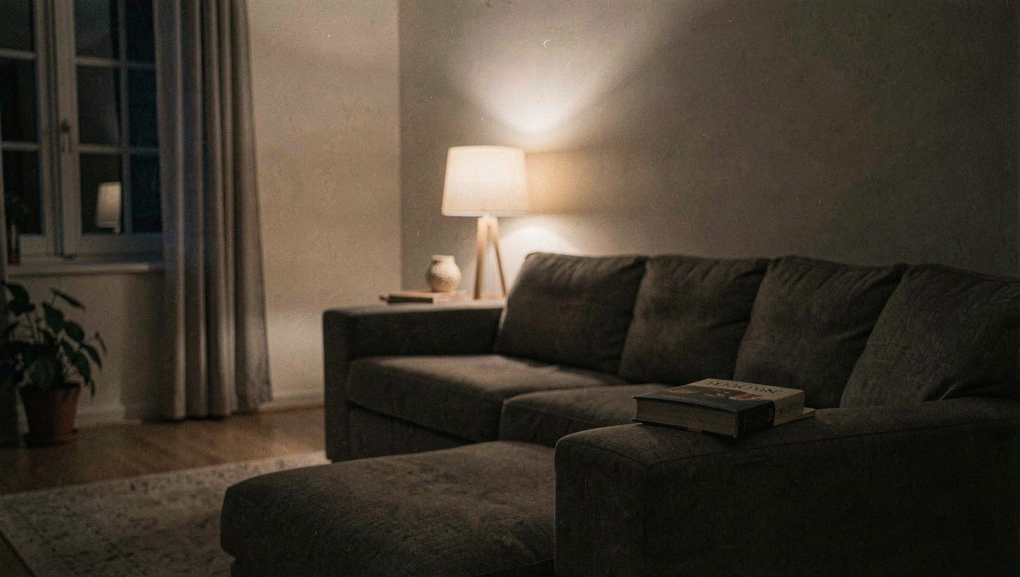 A living room after nine at night lit by one low table lamp, the ceiling light switched off, a book face down on the arm of a sofa