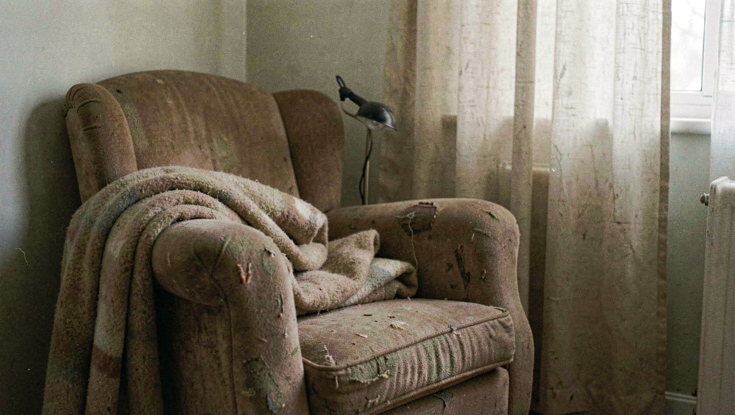A worn armchair in the corner of a room with a folded wool blanket over one arm and a small reading lamp behind it, curtains drawn