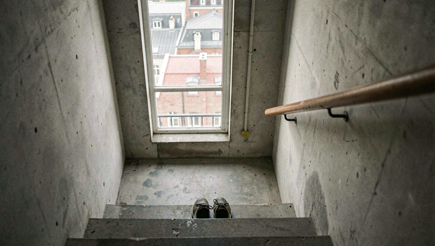 A concrete stairwell landing with a tall window looking onto rooftops, a pair of shoes at the top of the flight and a handrail catching the light