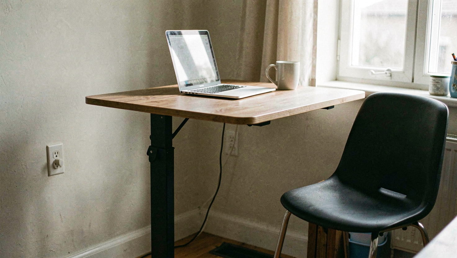A sit stand desk raised to standing height in a home office in the morning, a laptop and a mug on it, an empty chair to one side