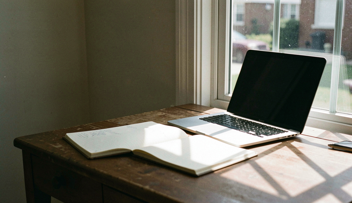 A desk pushed right up against a tall window in the afternoon, a laptop open on it and the shadow of the window bars falling across an open notebook